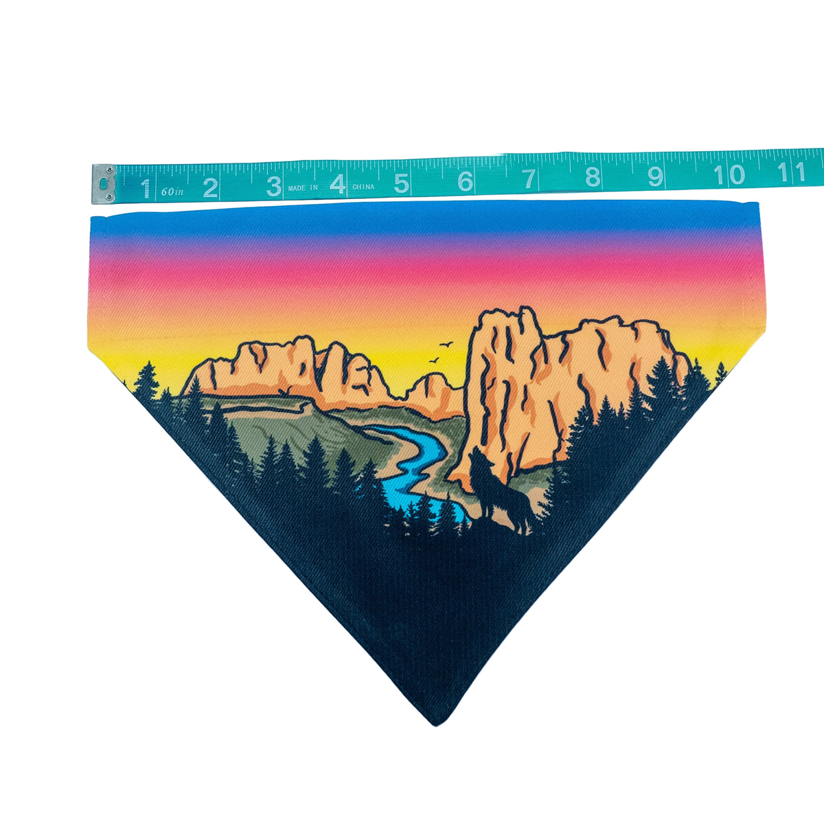 A triangular pet bandana featuring a colorful landscape print with a sunset gradient sky, mountains, trees, and a river, displayed on a white background with a measuring tape above it.